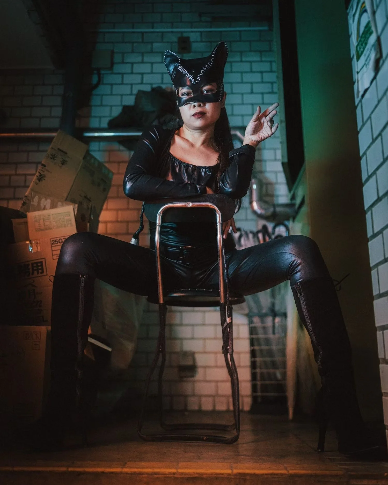 Person wearing a cat mask and black leather clothing sitting with legs spread apart in a dimly lit room, with cardboard boxes in the background.