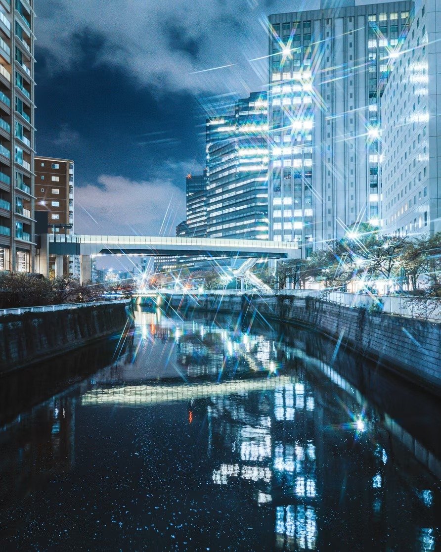 Night cityscape with illuminated modern buildings and a canal reflecting the city lights, enhanced with sparkling star effects.