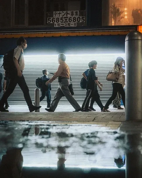 A group of pedestrians walking past a storefront with a glowing display, reflected in a puddle, from a Tokyo street photography workshop.