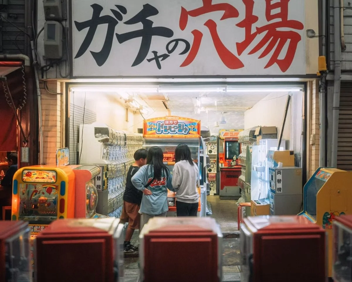 Three people gathered around a Japanese arcade game inside a small arcade shop, with numerous arcade machines and snack vending machines visible, and large Japanese signage above the entrance. Shot in Shinsekai, Osaka by Tokyo street photographer Den