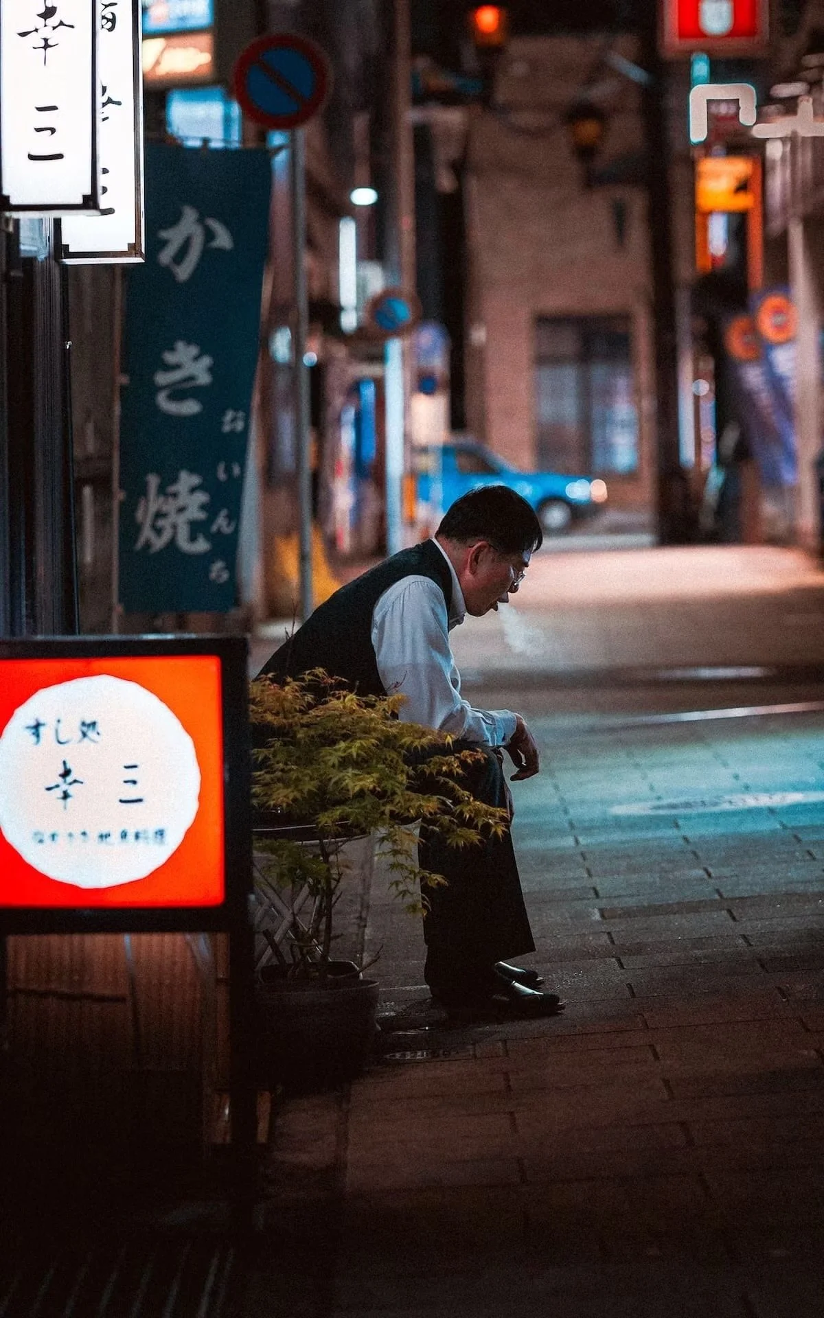 A man dressed in a white shirt and black vest sitting on a bench on a street at night, looking down, with a small plant nearby and illuminated signs in Japanese in the background.