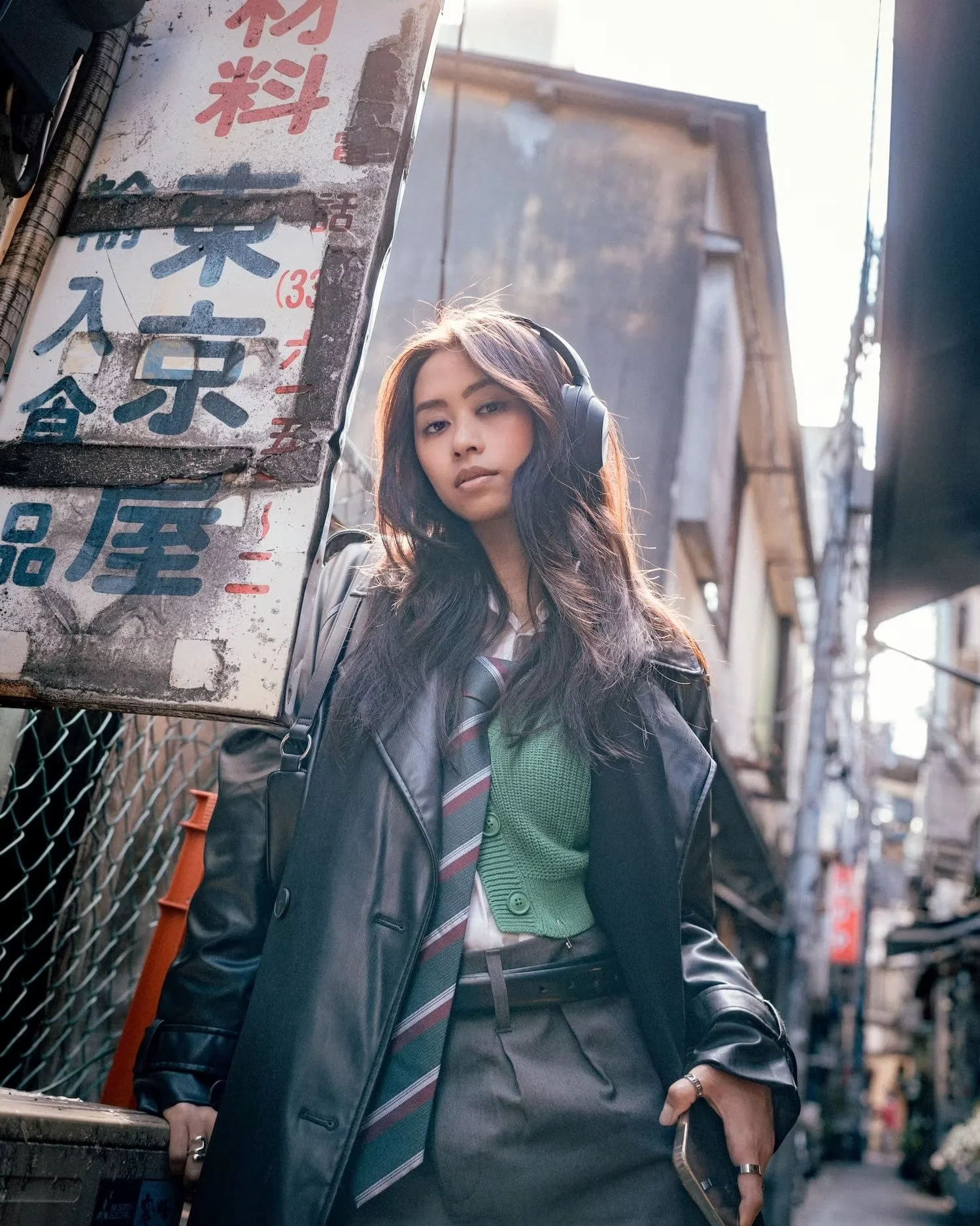 A young woman wearing headphones, a dark leather jacket, and a green sweater underneath, standing in an urban alleyway. She has long, wavy hair and is holding a smartphone in her right hand. Behind her are old buildings and a sign with Asian characte