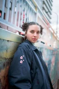A woman with curly hair and hoop earrings leans against a graffiti-covered fence. She is dressed in a dark jacket with Japanese characters and looks into the camera. The background shows an urban setting in Tokyo.