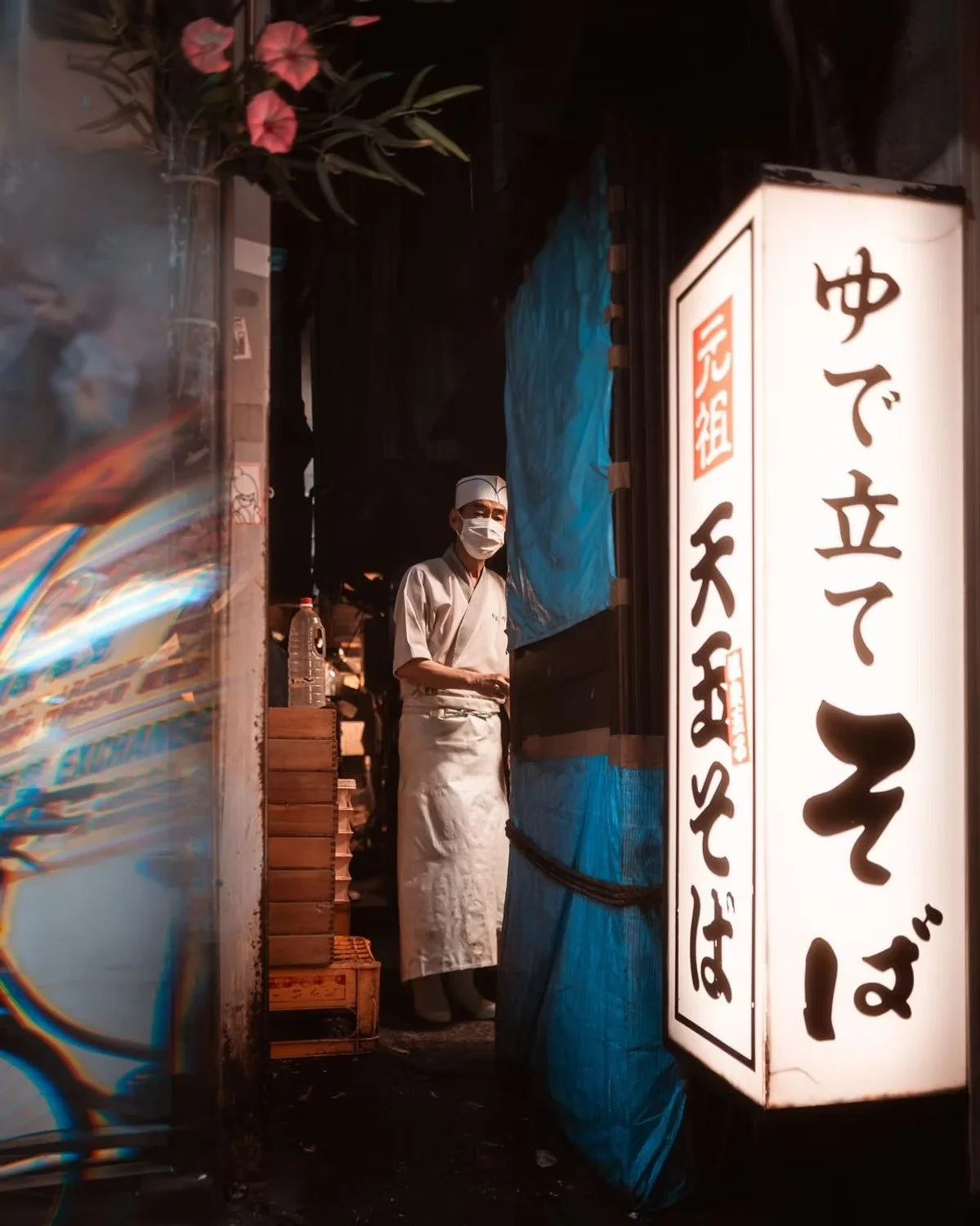 A chef wearing a face mask and white uniform standing inside a restaurant or food stall, with a large illuminated sign with Japanese writing beside him in Omoide Yokocho.