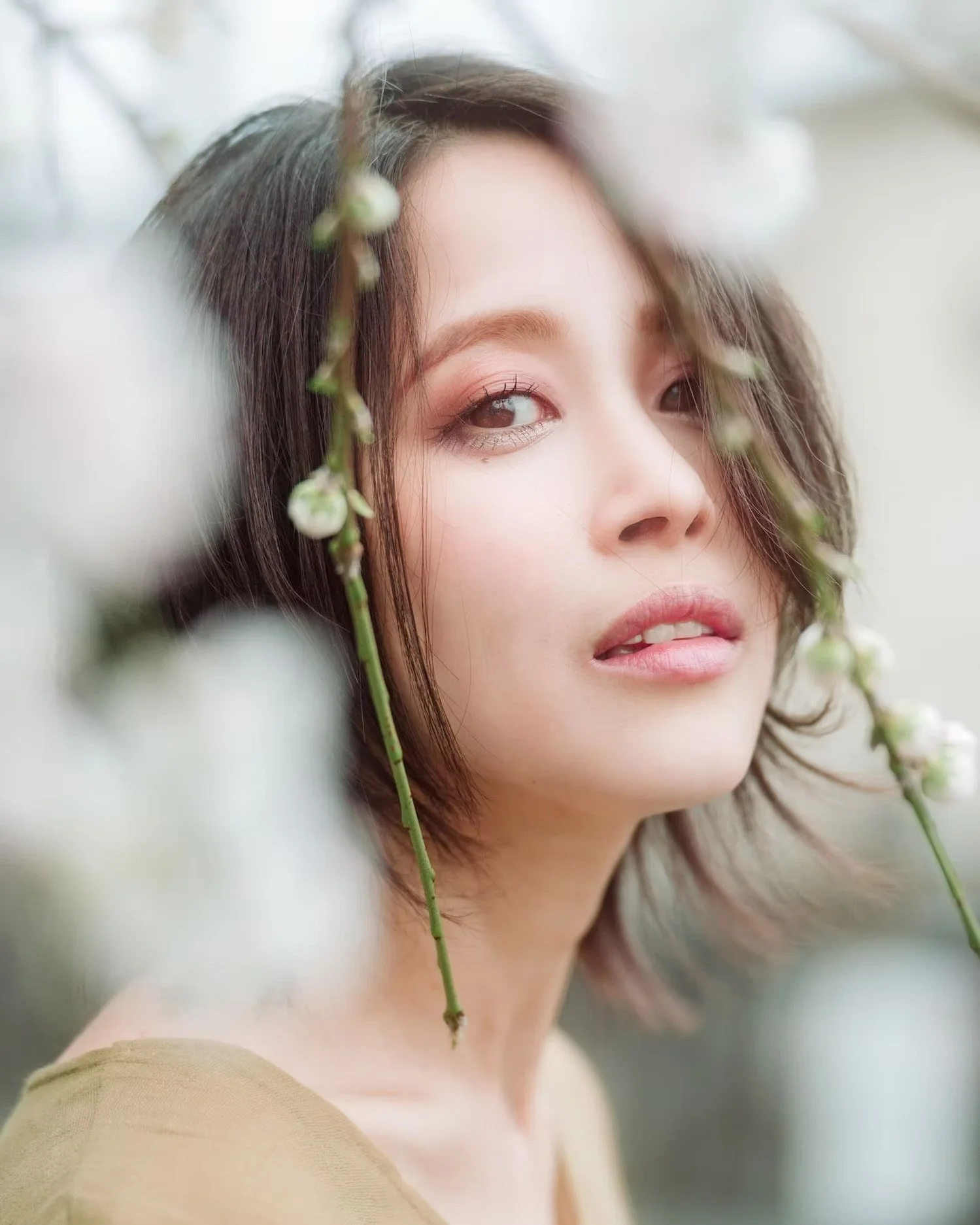 Close-up of a young woman with short brown hair and light makeup, partially obscured by white sakura cherry blossom flowers, looking at the camera. Shot by Tokyo portrait photographer Deniz Demir.
