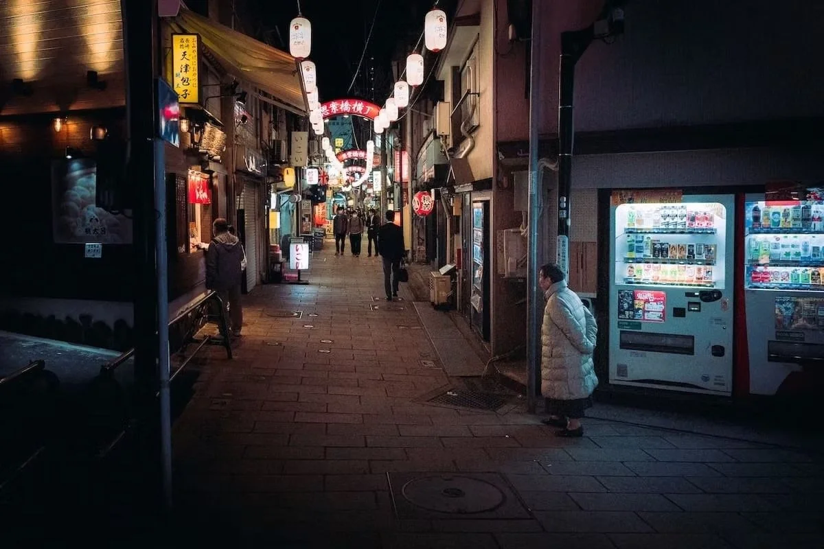 A narrow street at night with traditional Japanese lanterns hanging overhead. Several people are walking and talking, with some standing outside shops or vending machines. The street is dimly lit with signs and lights from small businesses. Nakagaki 
