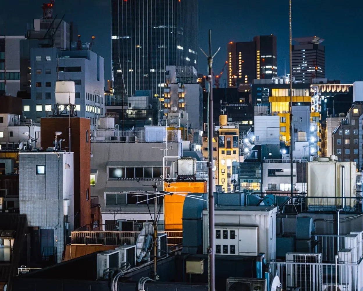 Nighttime cityscape of densely packed buildings with illuminated windows and various rooftop equipment, including antennas and air conditioning units, in an urban area.