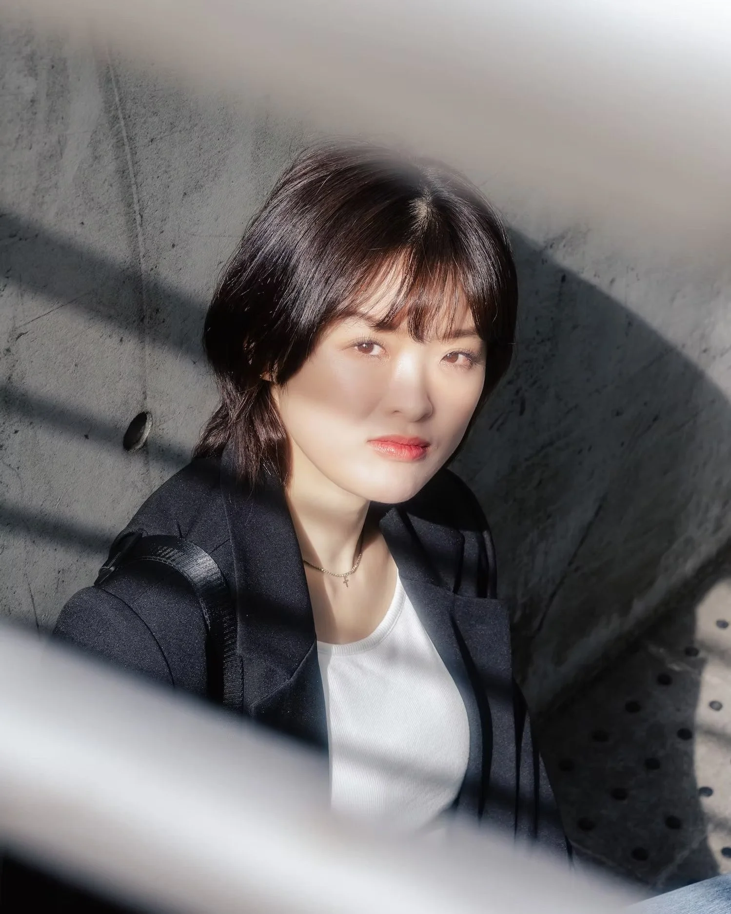 A woman with short dark hair, wearing a white shirt and black jacket, looking into the camera with a neutral expression, standing against a gray concrete wall.