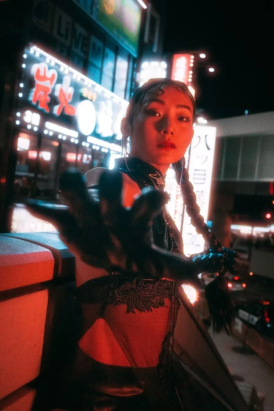 A woman with braided hair and earrings extending her hand towards the camera in a city at night, with illuminated signs in Japanese characters in the background.