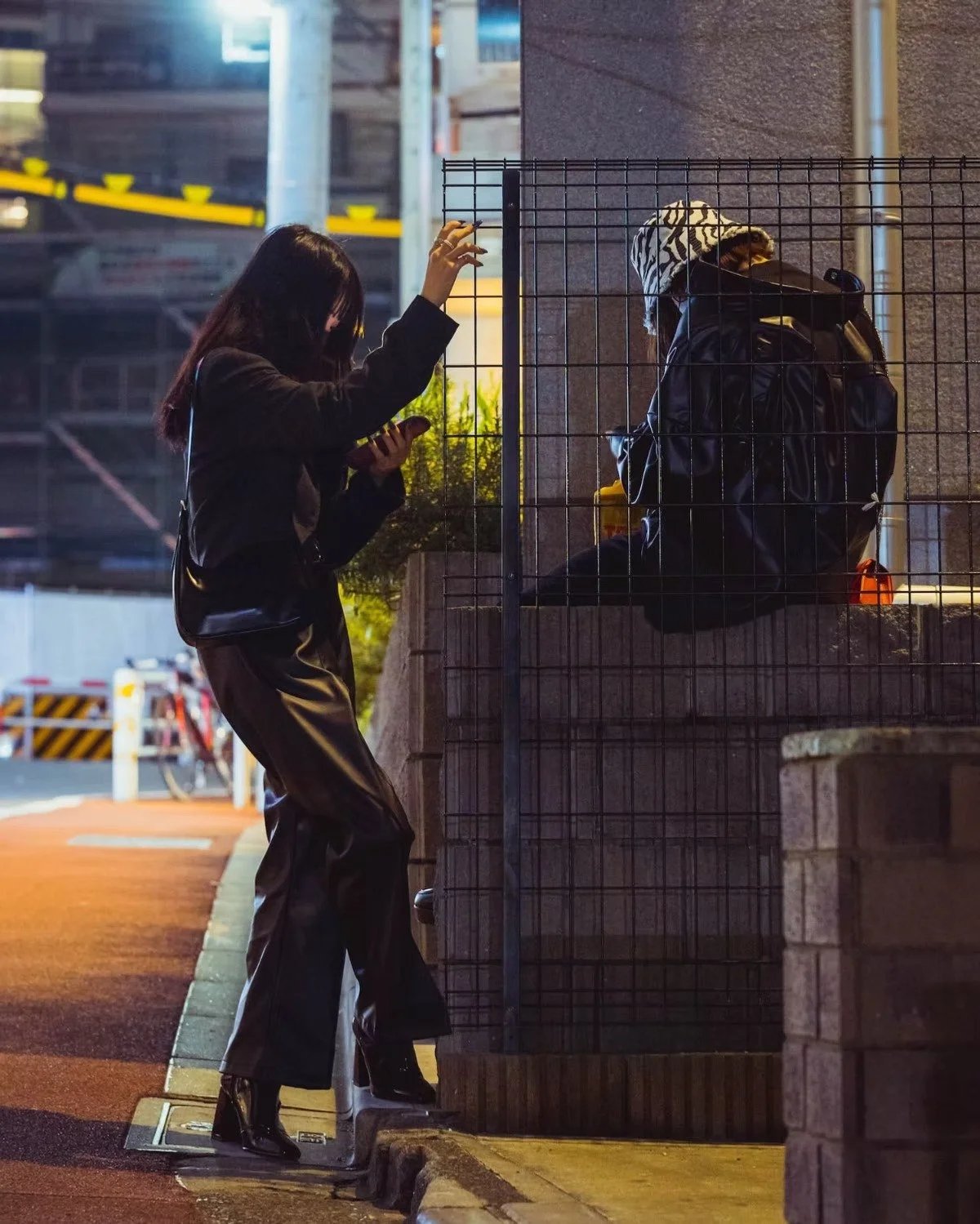 A woman standing on a sidewalk, looking at her phone, in front of a seated person behind a metal gate at night with her friend in Shibuya.