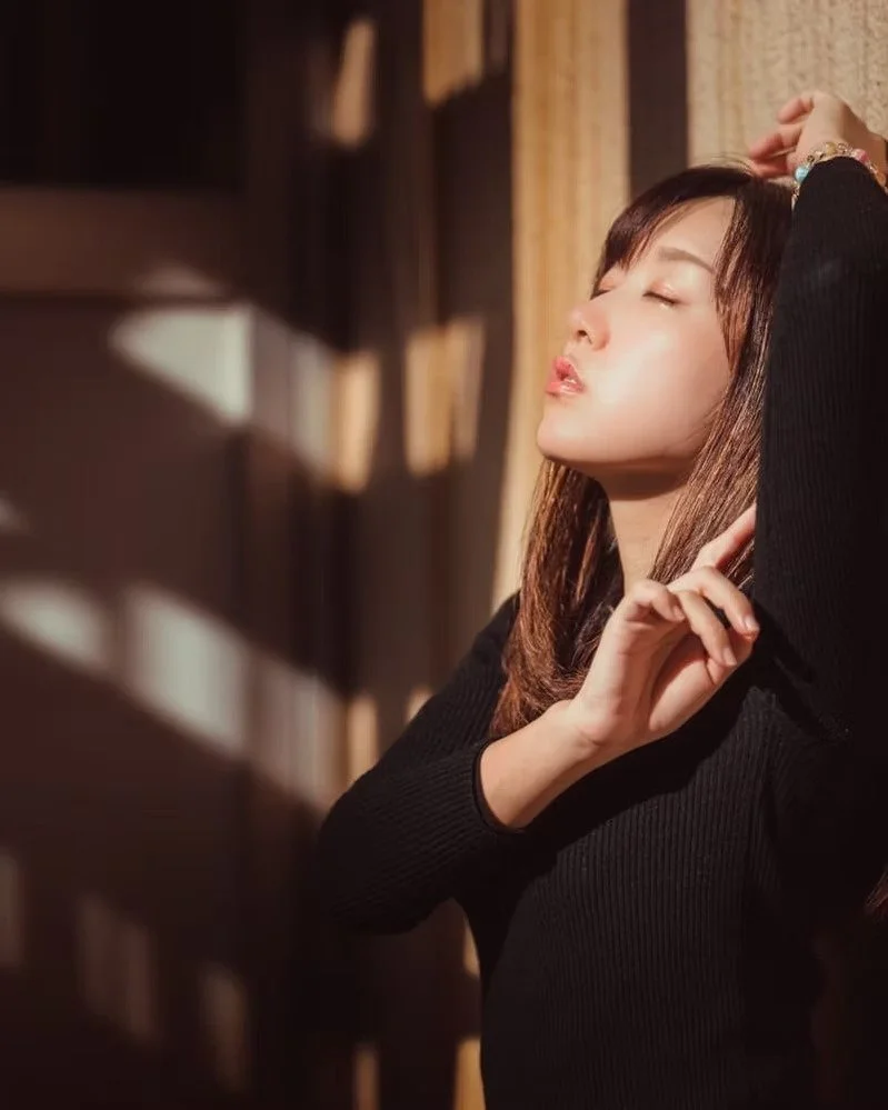 A young woman with closed eyes relaxing in sunlight, standing near a wooden wall with shadows cast by blinds.