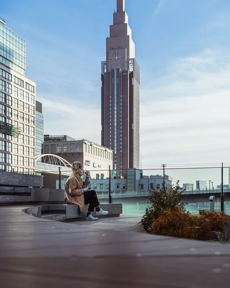 A person sitting on a rooftop bench, drinking from a cup, with a tall skyscraper and city buildings in the background during daytime.