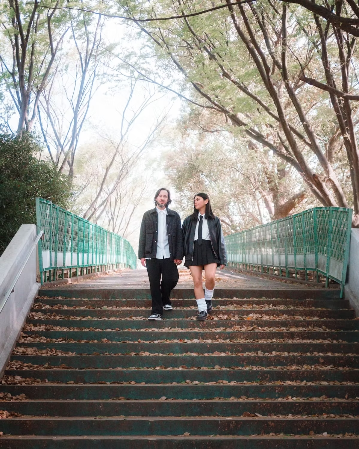 A couple walks hand-in-hand down a flight of stairs in a park, surrounded by trees with autumn leaves. The sunlight filters through the trees, creating a peaceful and romantic atmosphere.