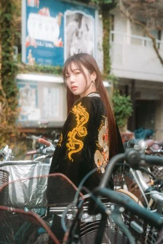 Young woman with long brown hair standing among bicycles on a city street, wearing a black jacket with gold dragon embroidery.