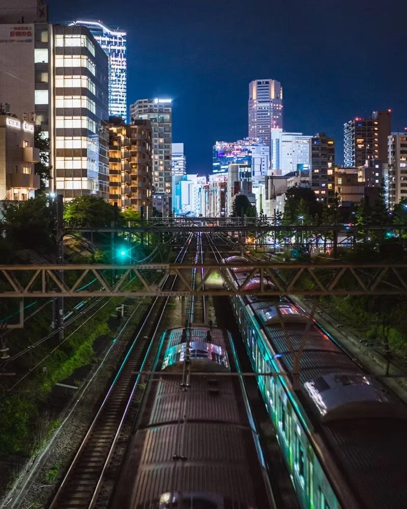 Nighttime cityscape with tall illuminated buildings, train tracks in the foreground, and two green and silver trains moving on the tracks.
