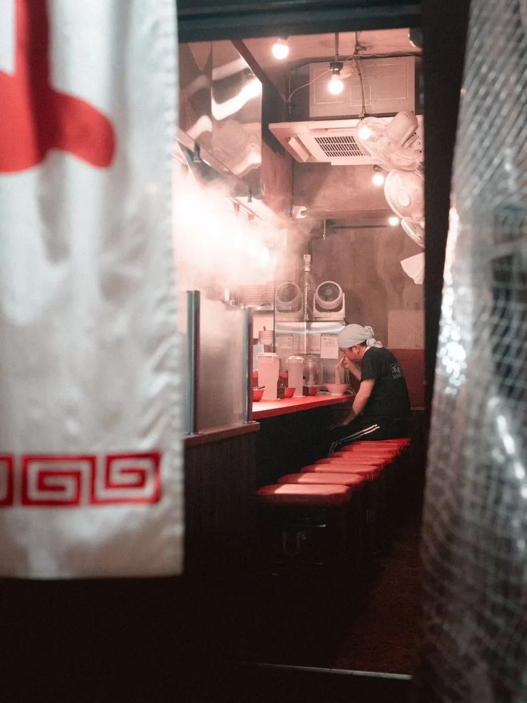 A person sitting at a counter in a small restaurant, wearing a white hair covering and a black shirt, with a bowl in front of them, surrounded by kitchen equipment and ventilation fans.