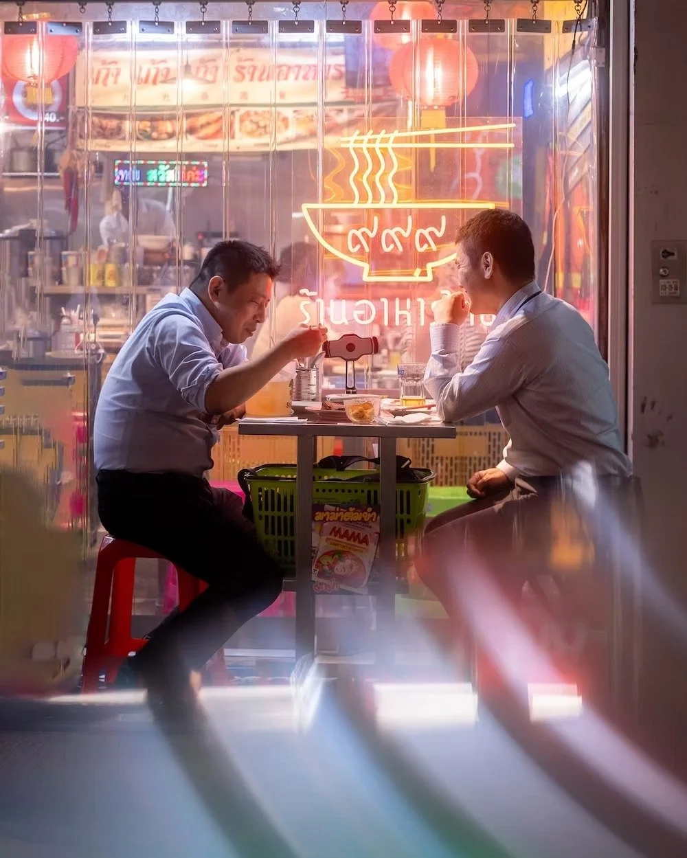 Two men sit at a small table in a Thai restaurant, eating noodles. The restaurant has a neon sign of a steaming bowl of noodles behind them, and a glass window reflecting the street outside. Shimbashi Tokyo.