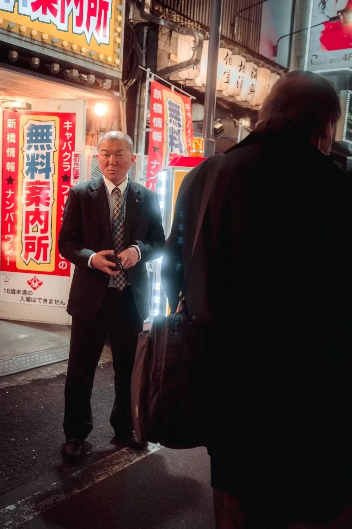 A man in a suit and tie standing on a city street at night, holding a phone, with illuminated signs and banners in Japanese behind him, and another person partially visible in the foreground in Shimbashi Tokyo.