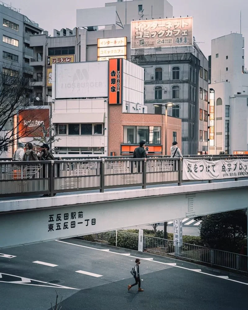 City scene with pedestrians walking on an elevated sidewalk, Japanese signs on buildings, and a person crossing the street below.