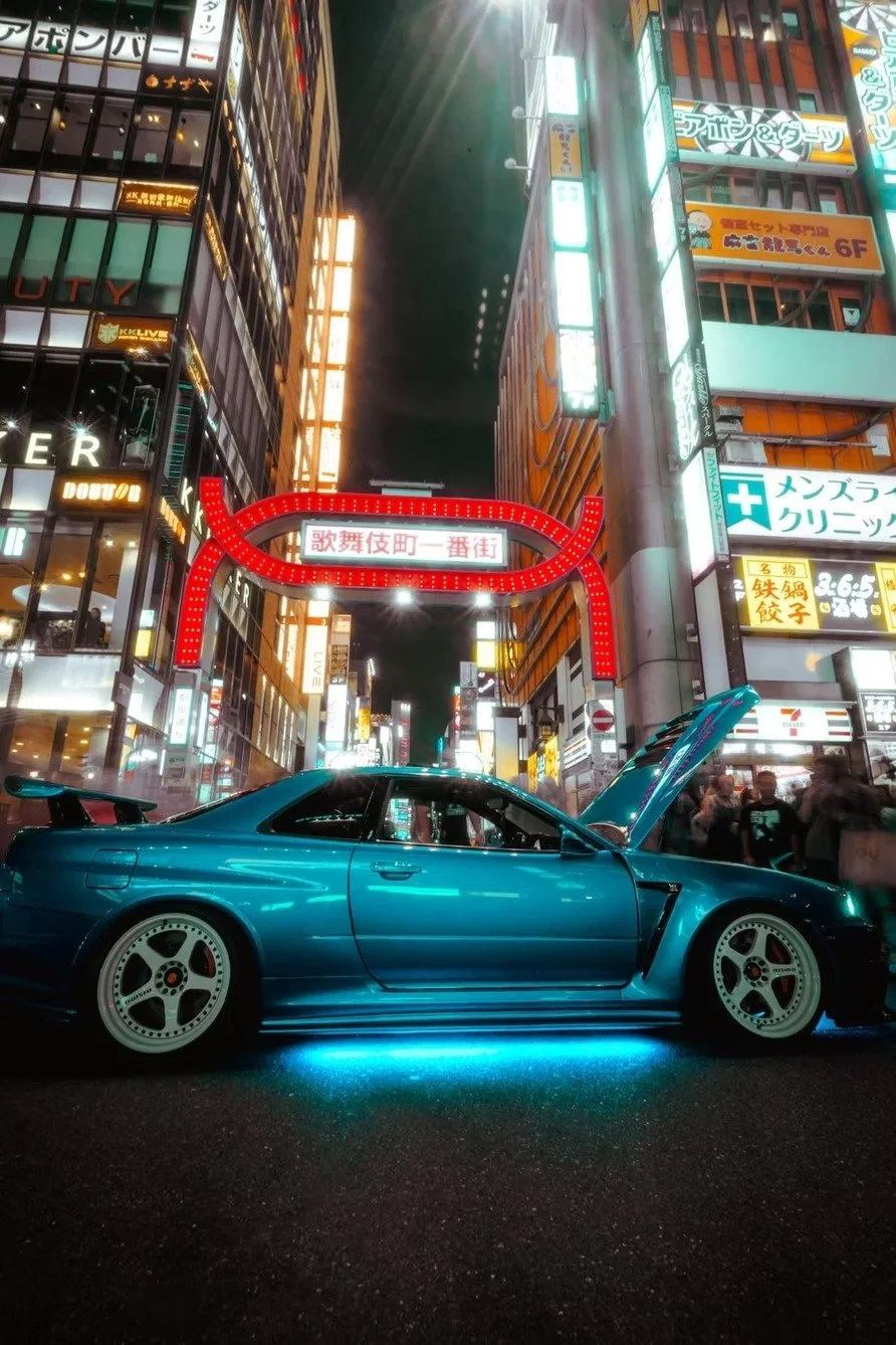 A blue sports car with its hood open parked on a city street illuminated by bright neon signs at night.