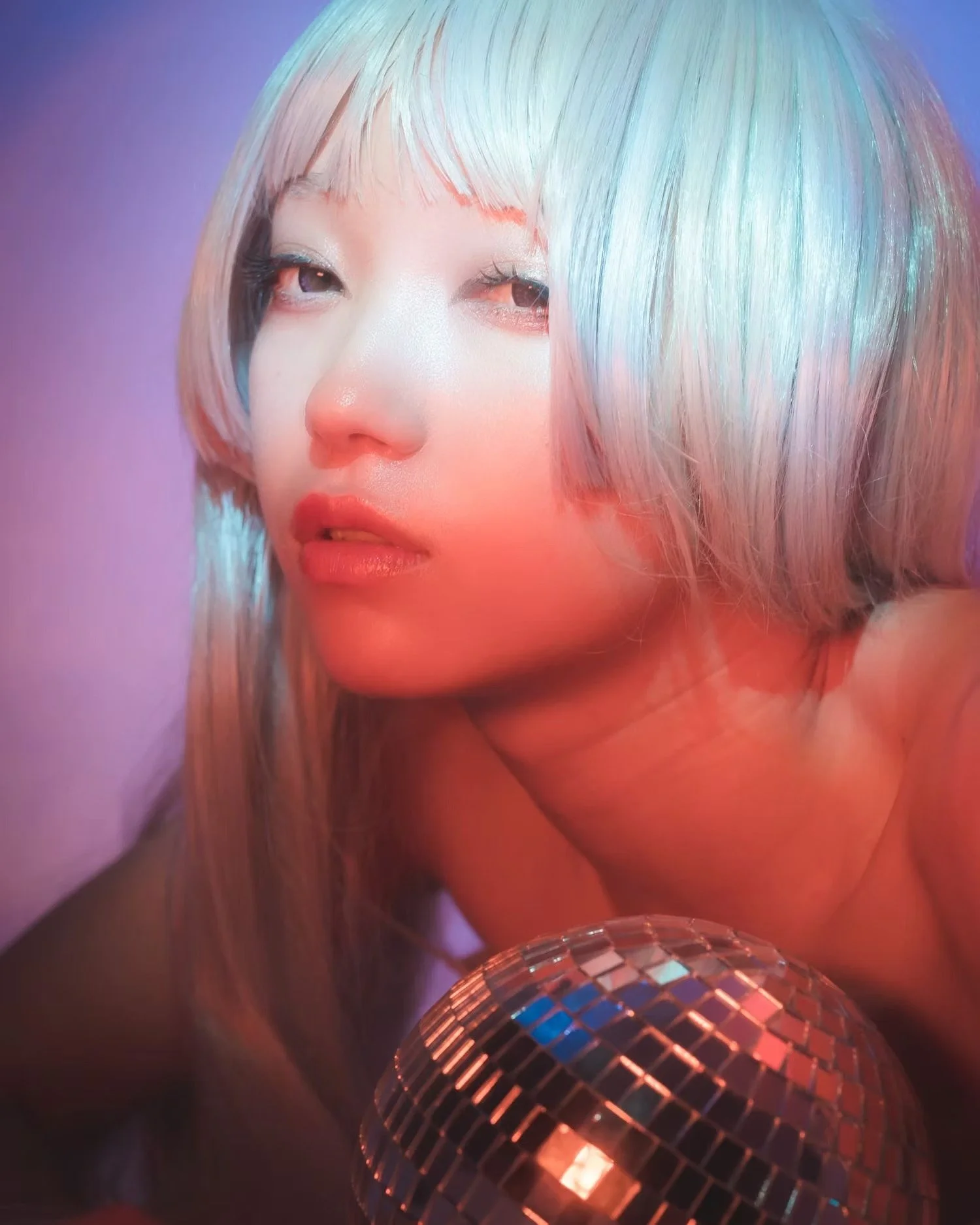 A person with multi-colored hair and makeup, holding a disco ball, with pink and purple lighting.