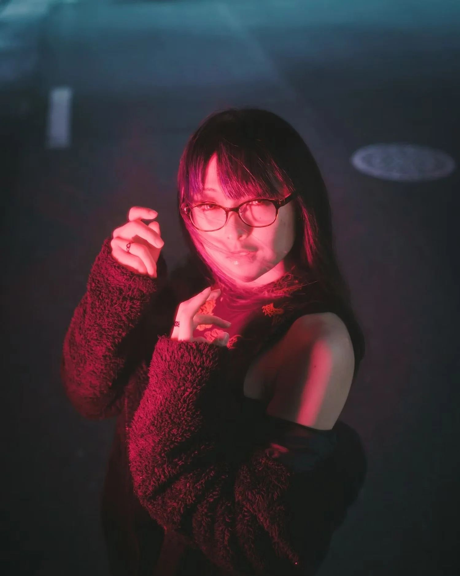 A young woman with dark hair and glasses, wearing a black sleeveless top and a fuzzy red long-sleeve sweater, striking a pose with her hand near her face in a dark environment with pink and blue lighting.