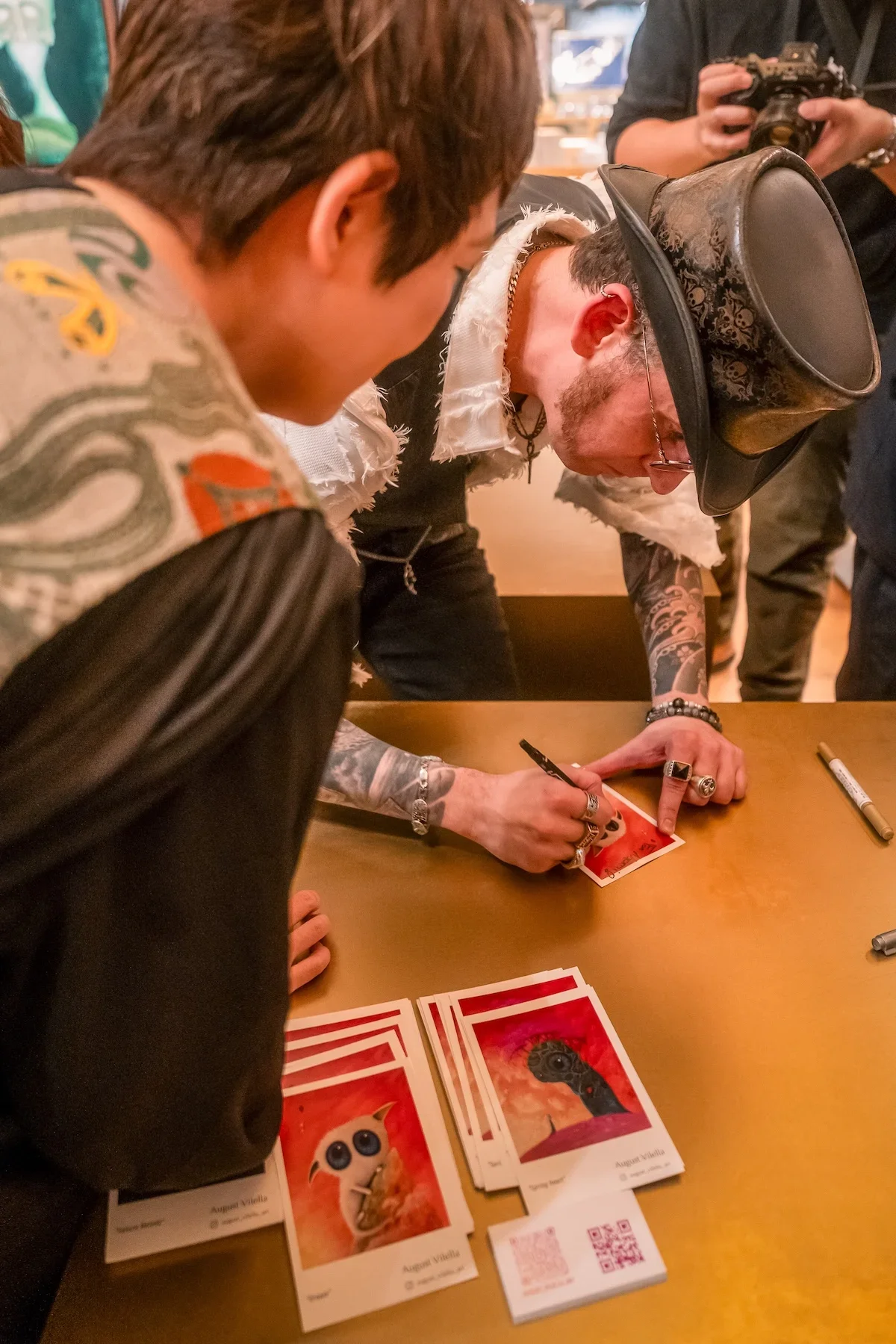 August Vilella
---
Close-up photograph of Spanish surrealist artist August Vilella wearing a black hat and round glasses as he signs a colourful art print at his Tokyo exhibition. His tattooed arms and hands are visible as he personalizes the artwork