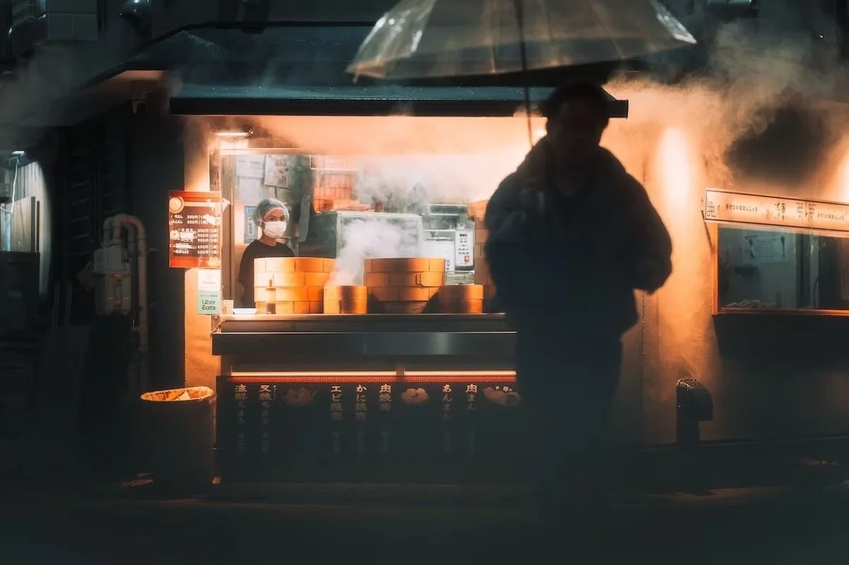A street food stand at night with steam rising from cooking. A person wearing a face mask stands behind the counter, and another person is silhouetted under an umbrella in front of the stand. Nakano at night.
