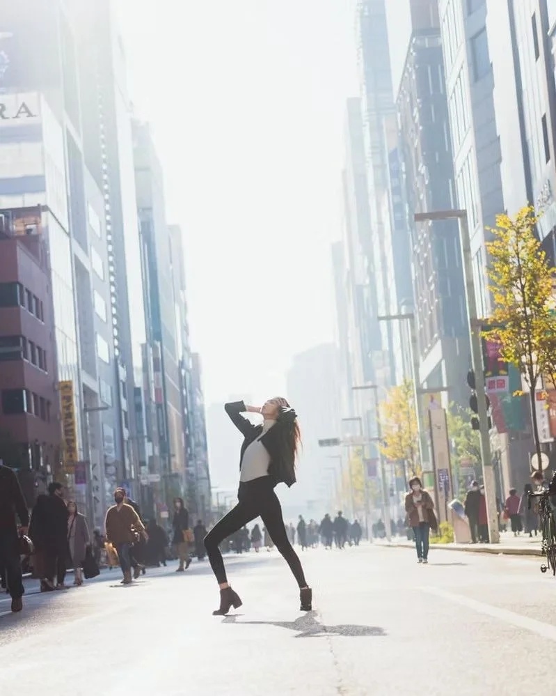 A woman dancing on a city street with skyscrapers, people walking, and sunlight shining brightly. Shot by Tokyo portrait photographer Deniz Demir.