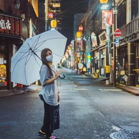 A woman in a mask holds a white umbrella on a wet Tokyo street, neon lights reflecting off the pavement as lanterns line the alleyway.