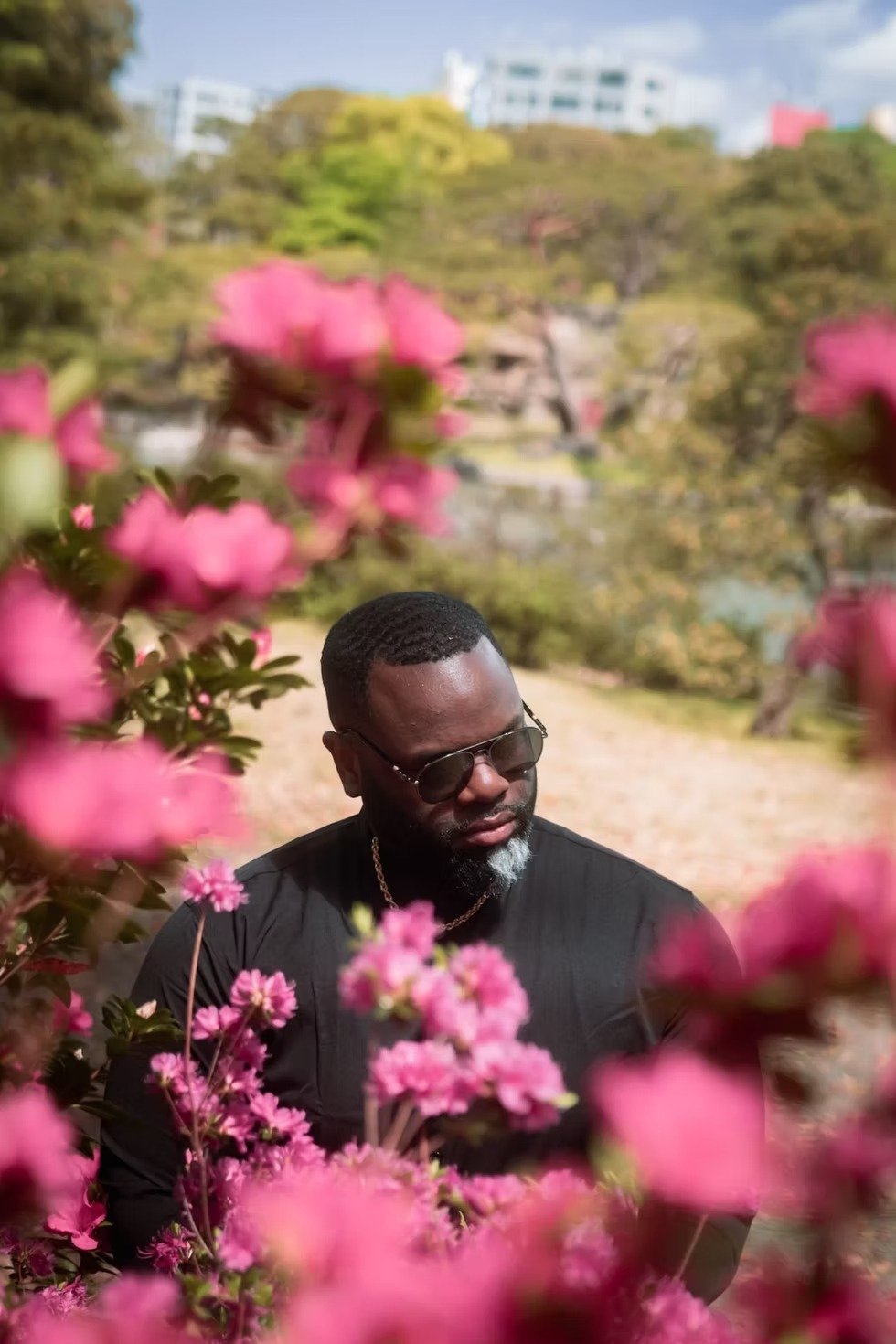 A man in a black shirt stands in a park surrounded by blooming pink flowers. The man gazes thoughtfully as he enjoys the peaceful nature setting.