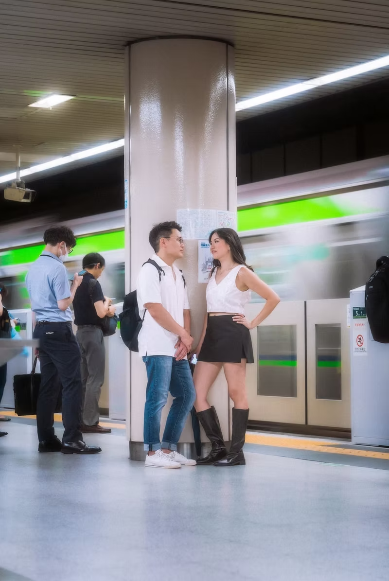 A group of people is seen at a Tokyo train station. Two individuals are talking to each other while others wait at the platform. The clean, modern architecture of the station is visible in the background.