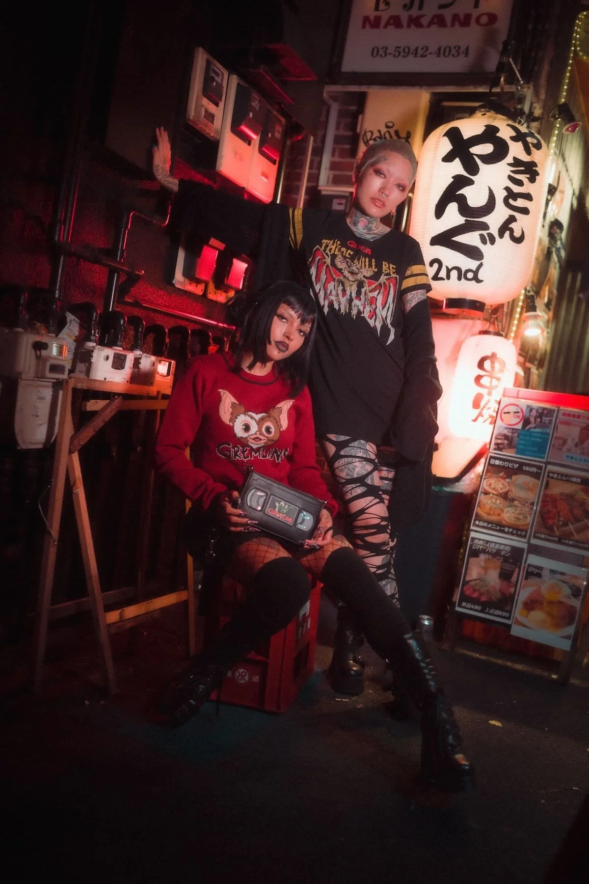 Two women with alternative fashion styles posing in a dimly lit, Japanese-themed restaurant or bar. One is sitting on a stool holding a video game controller, wearing a red Gremlins sweater, fishnet stockings, and black boots. The other stands beside her, wearing a black graphic shirt, ripped pants, and has numerous tattoos and piercings. Large Japanese lanterns and menu boards are visible in the background.