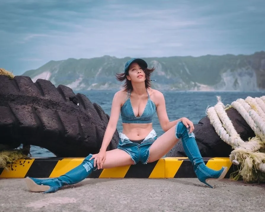 Young woman in denim outfit sitting on a yellow and black barrier near the water with cliffs in the background, posing with legs spread and hands on the ground.