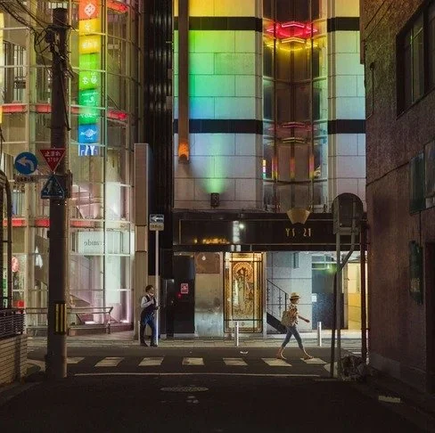 A city street at night with a rainbow-lit building reflecting colors, as pedestrians walk and cross the road, adding movement to the urban scene.