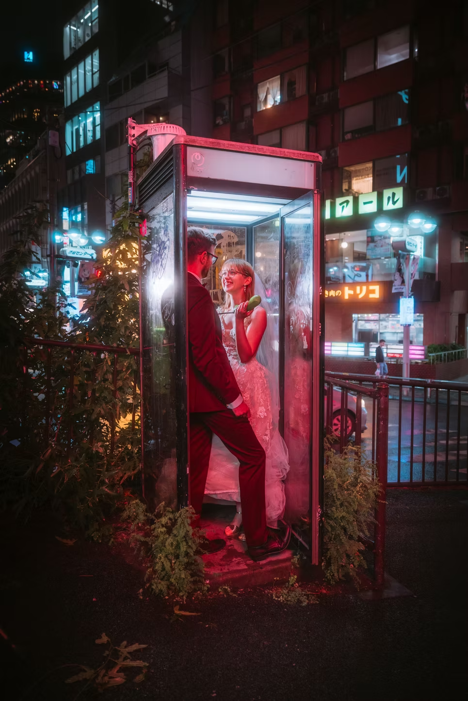A couple dressed for a wedding standing inside a lit phone booth on a city street at night, smiling at each other. The scene is illuminated by neon lights and signs.