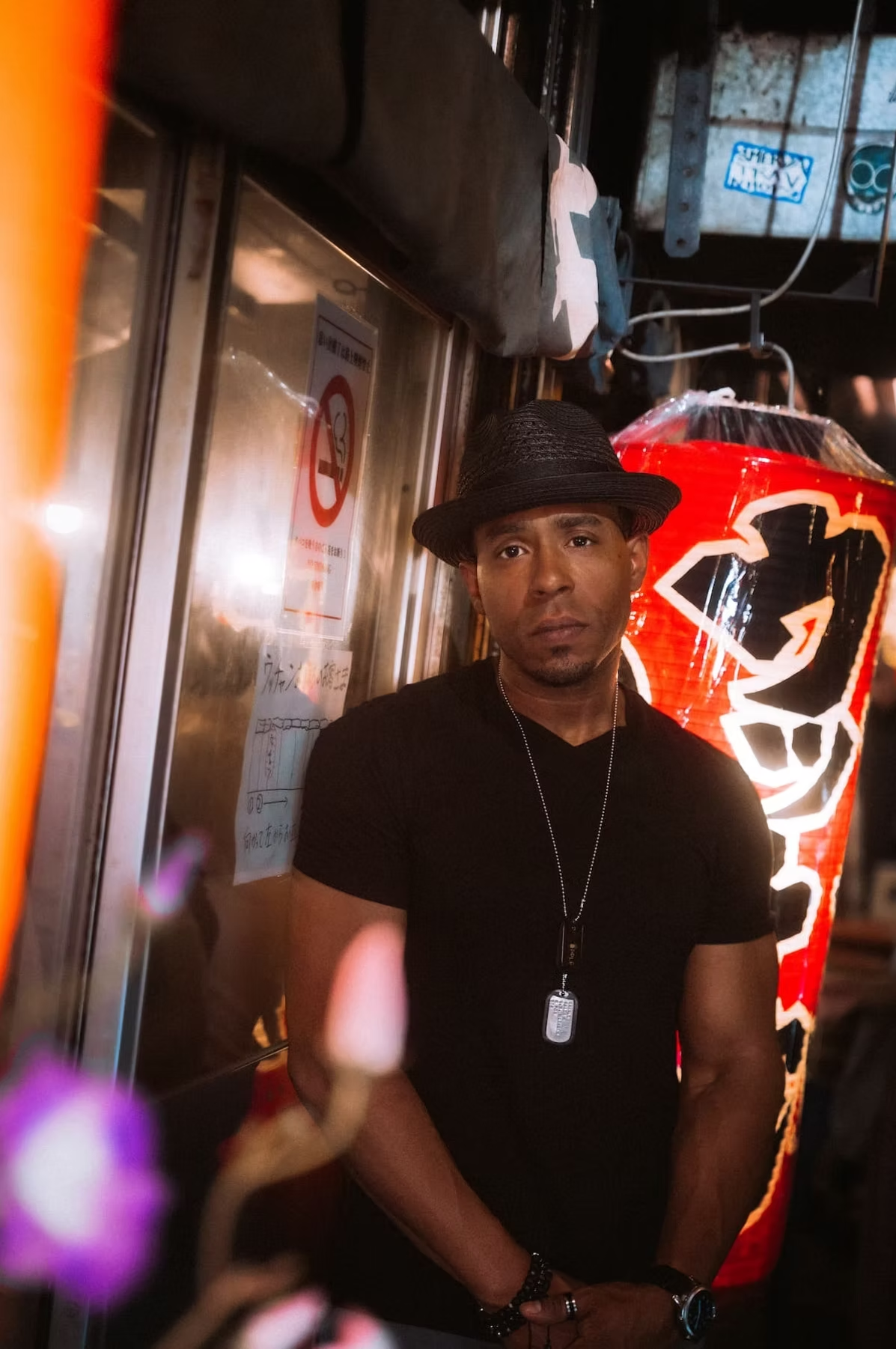A man in a black shirt and hat stands in front of a glowing red Japanese lantern and neon signs in a busy Tokyo alley, giving a serious expression.