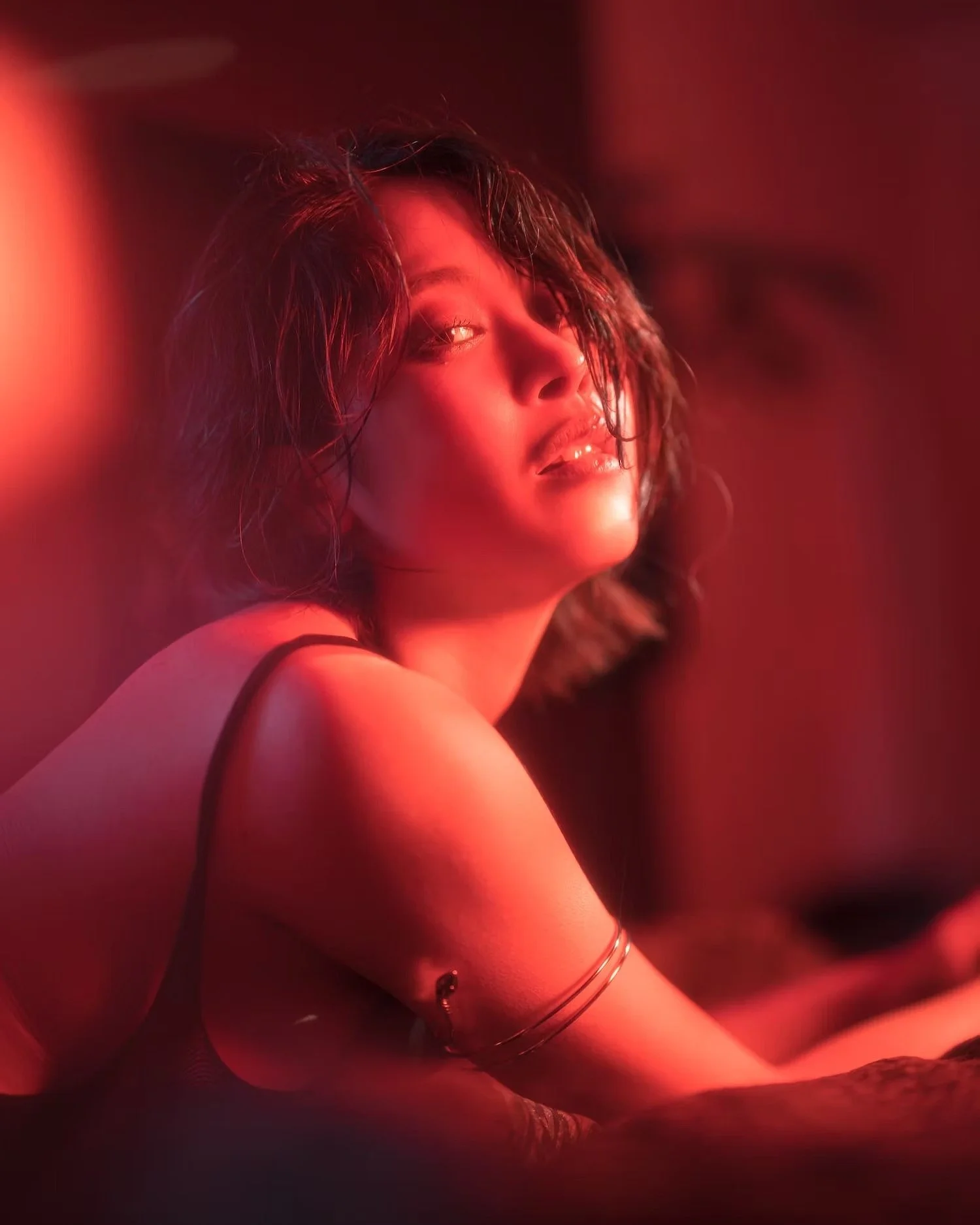 A woman with dark wavy hair illuminated by red lighting, wearing a black strappy top and silver jewelry, looking over her shoulder with a sultry expression.