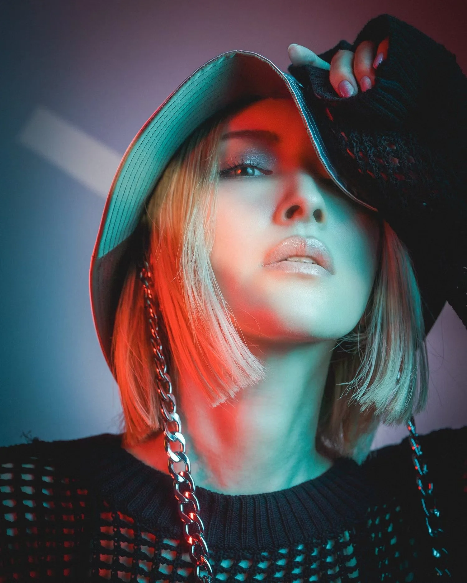 Close-up of a woman with blonde hair wearing a black fishnet top, chunky chain necklace, and a wide-brimmed hat, illuminated by colorful neon lighting.