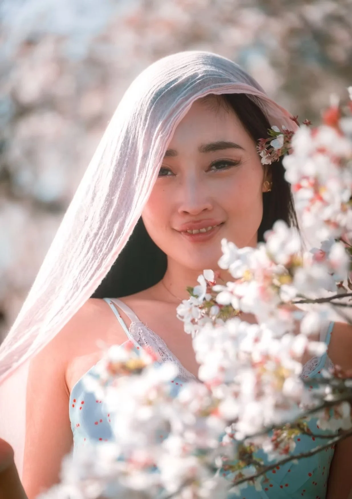 A woman stands in a cherry blossom garden, smiling gently while wearing a sheer veil. The delicate flowers frame her face, creating a serene and peaceful atmosphere.