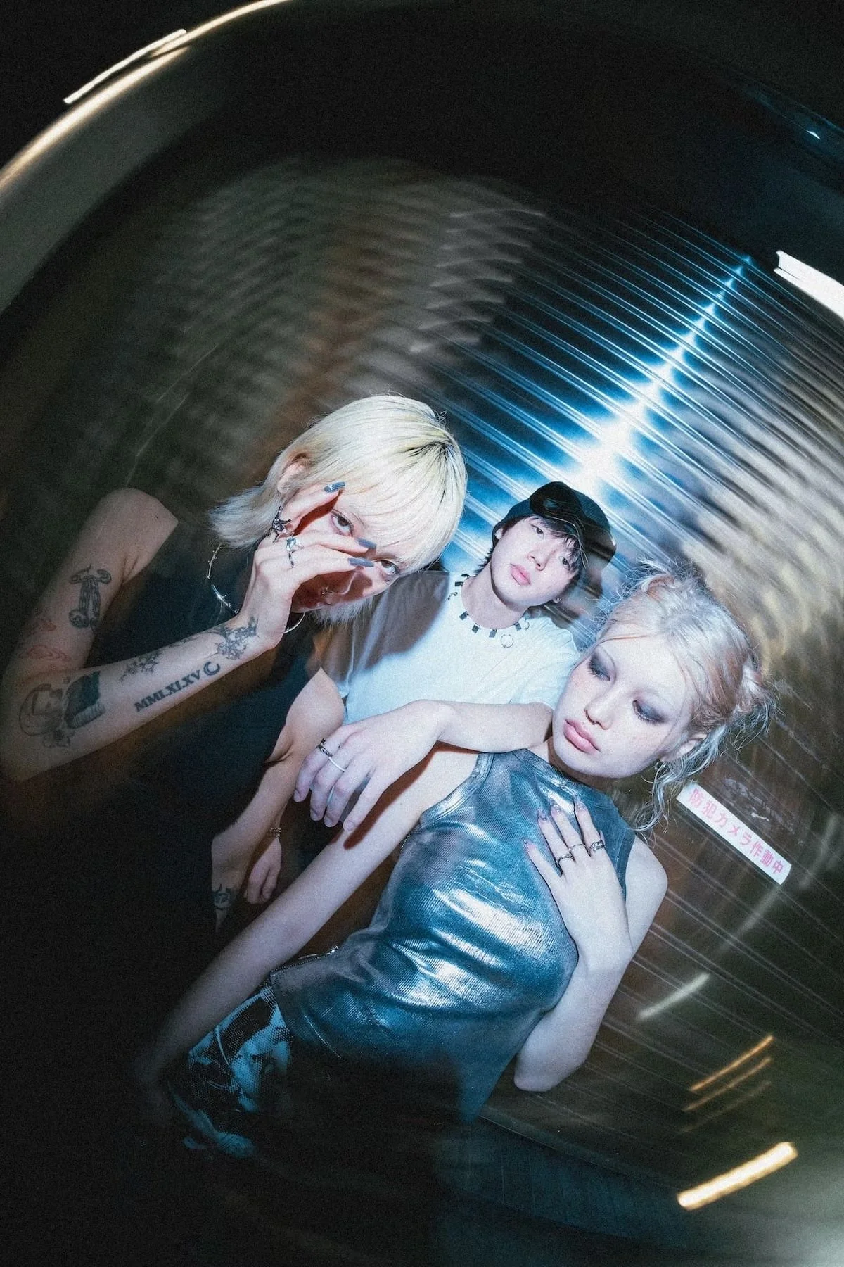 Three young women with styled hair and trendy clothing posing together in a dark, metallic environment with blue and white lighting.
