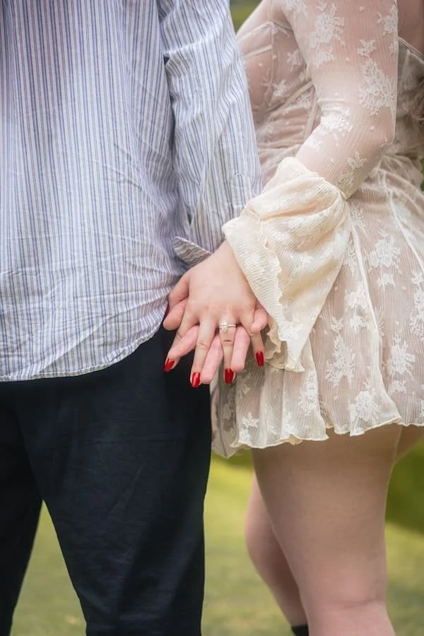 A man and woman romantically holding hands outdoors, showcasing their wedding ring with the woman wearing a lace dress and the man in a striped shirt and dark pants shot by couples tokyo photographer Deniz Demir.