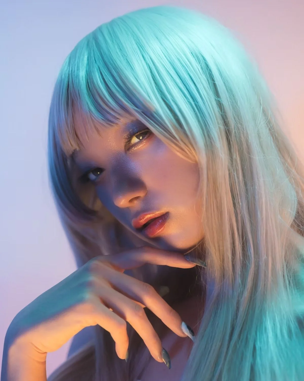 A young woman with platinum blonde hair with blue streaks, posing with her hand near her face in a colorful, artistic lighting setting.