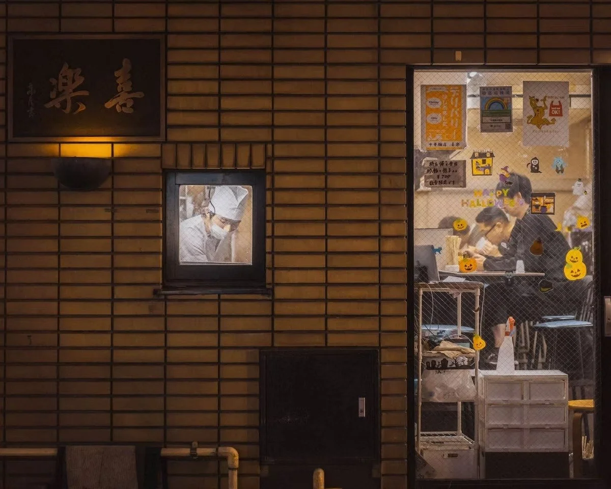 Nighttime view of a brick restaurant with a small window showing a chef wearing a white hat and face mask. The interior behind the glass door is decorated with Halloween-themed stickers and posters, and people are working inside.