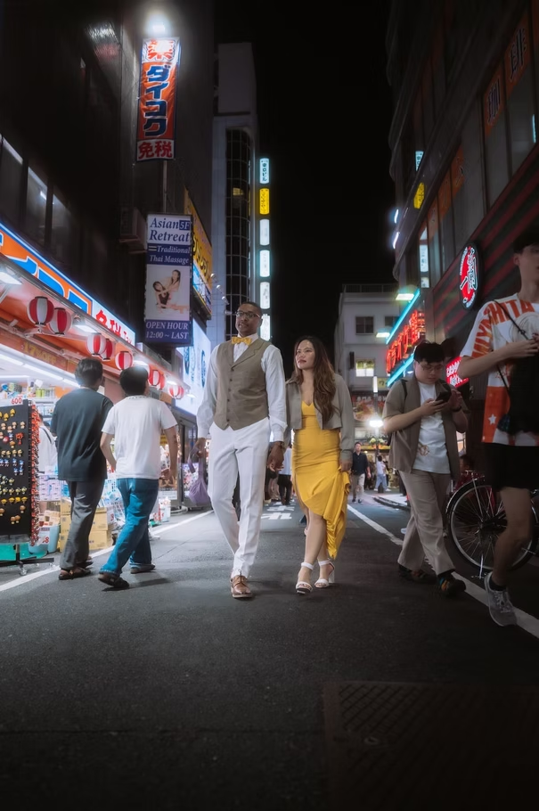 People walking on a busy city street at night, illuminated by bright signs and storefronts.