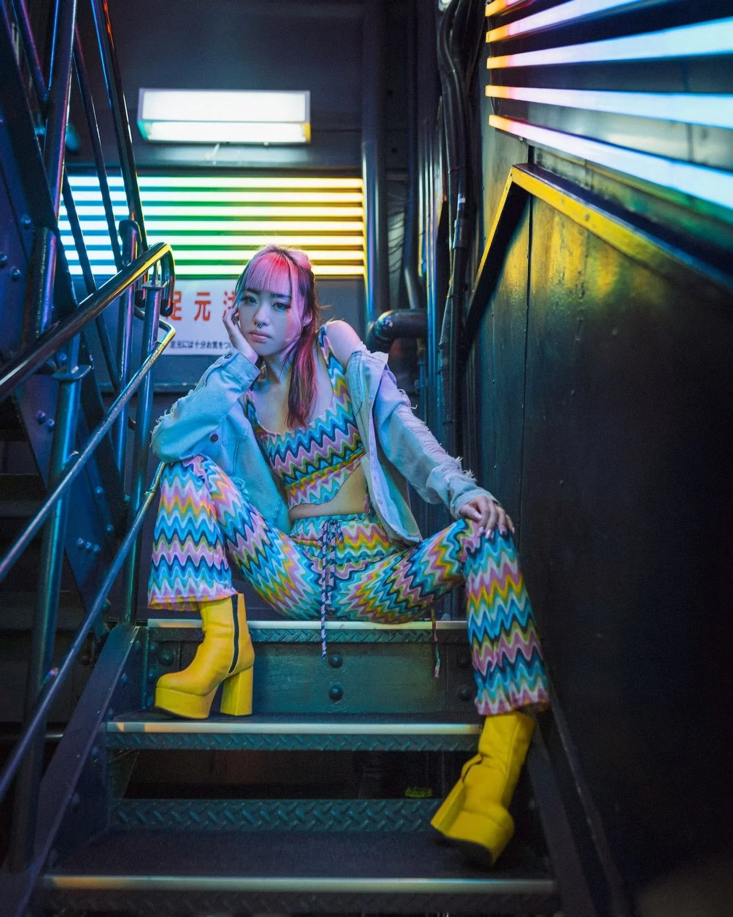 A young woman with pink hair sitting on metal stairs in front of neon lights, wearing colorful zigzag-patterned pants, yellow rain boots, and a light jacket.