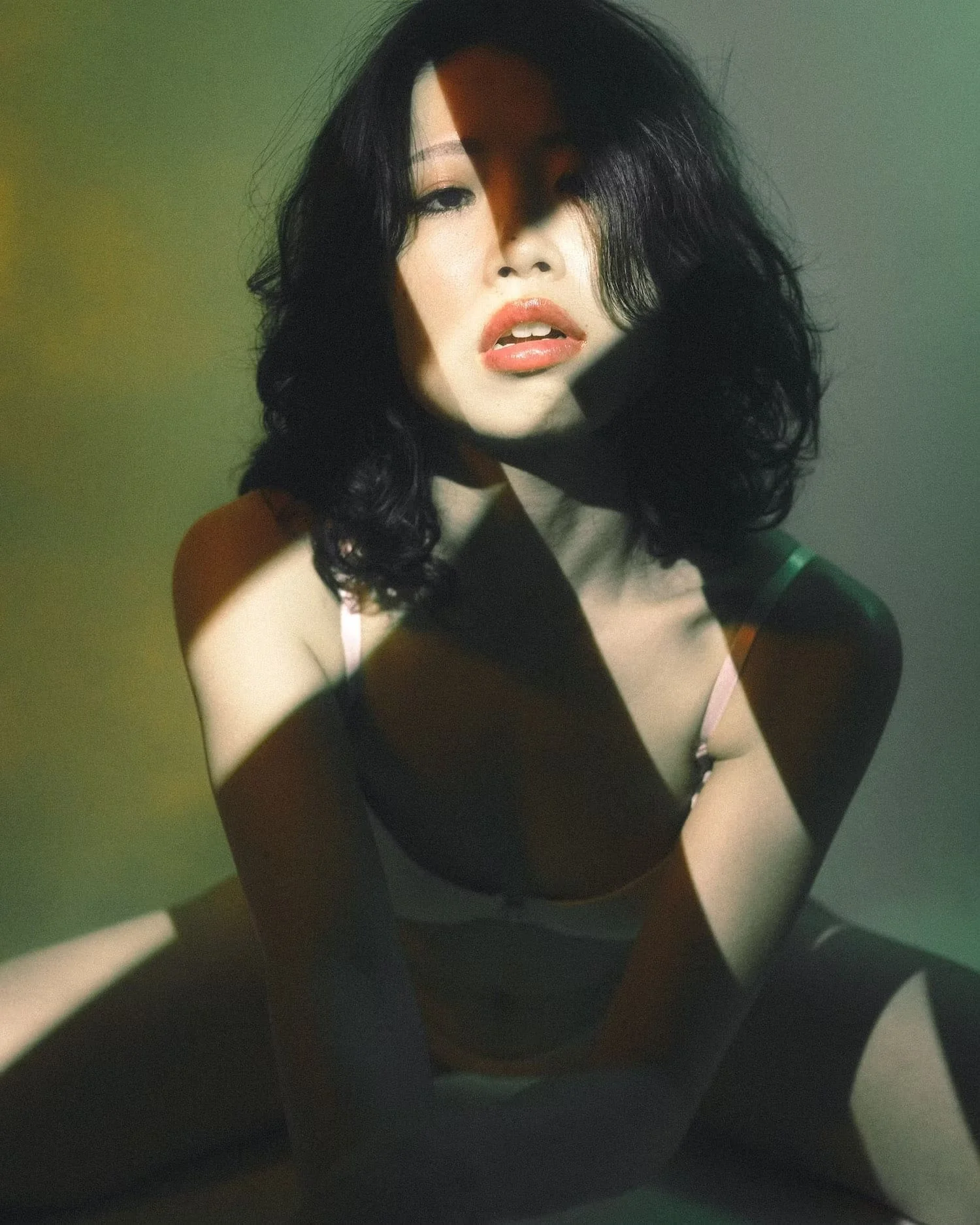 A woman with black, curly hair and light skin tone, wearing a black top with spaghetti straps, looking directly at the camera. She is partially obscured by shadows and the photo has a dark, moody ambiance.