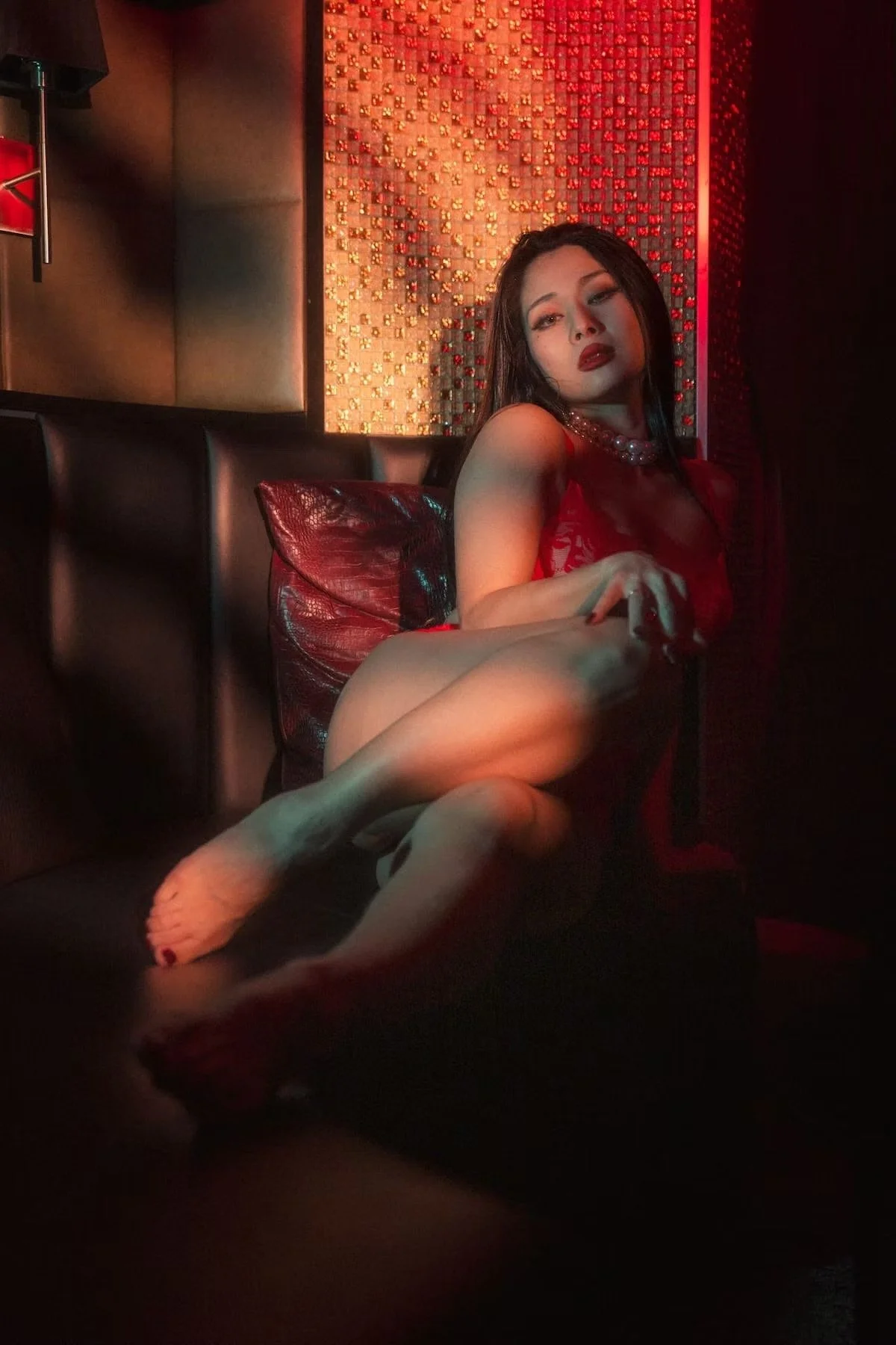 A woman with dark hair and red lipstick sitting on a dark couch, wearing a red dress and pearl necklace, with a red leather pillow and a textured red and gold wall behind her.