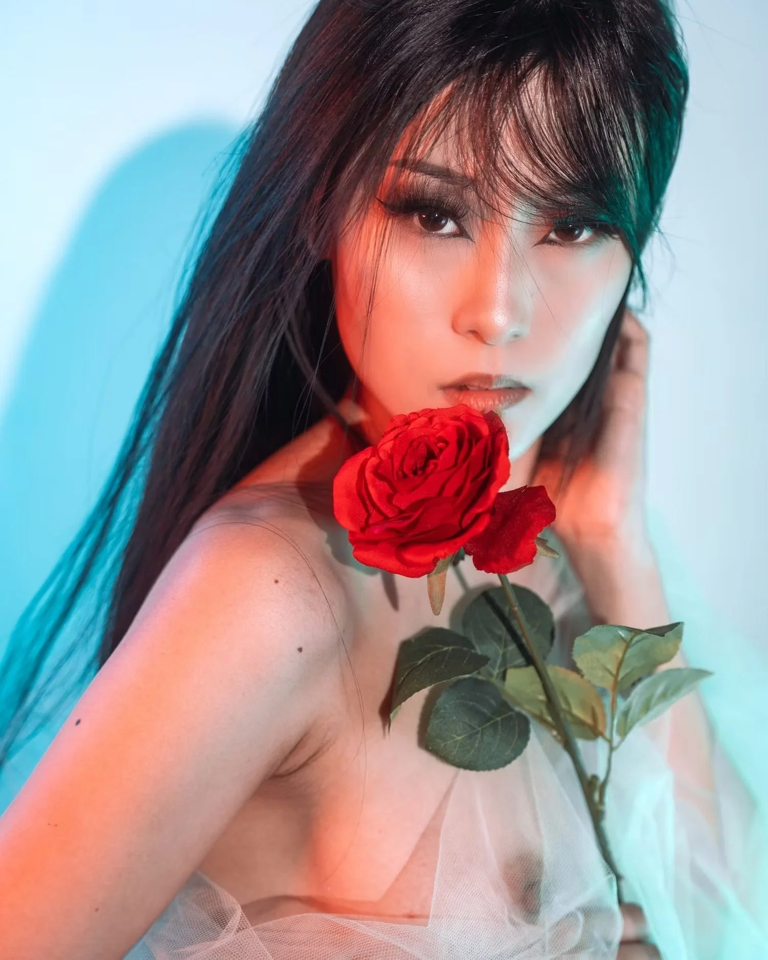 A woman with long dark hair holding a red rose near her face, with a soft expression and a gentle gaze.