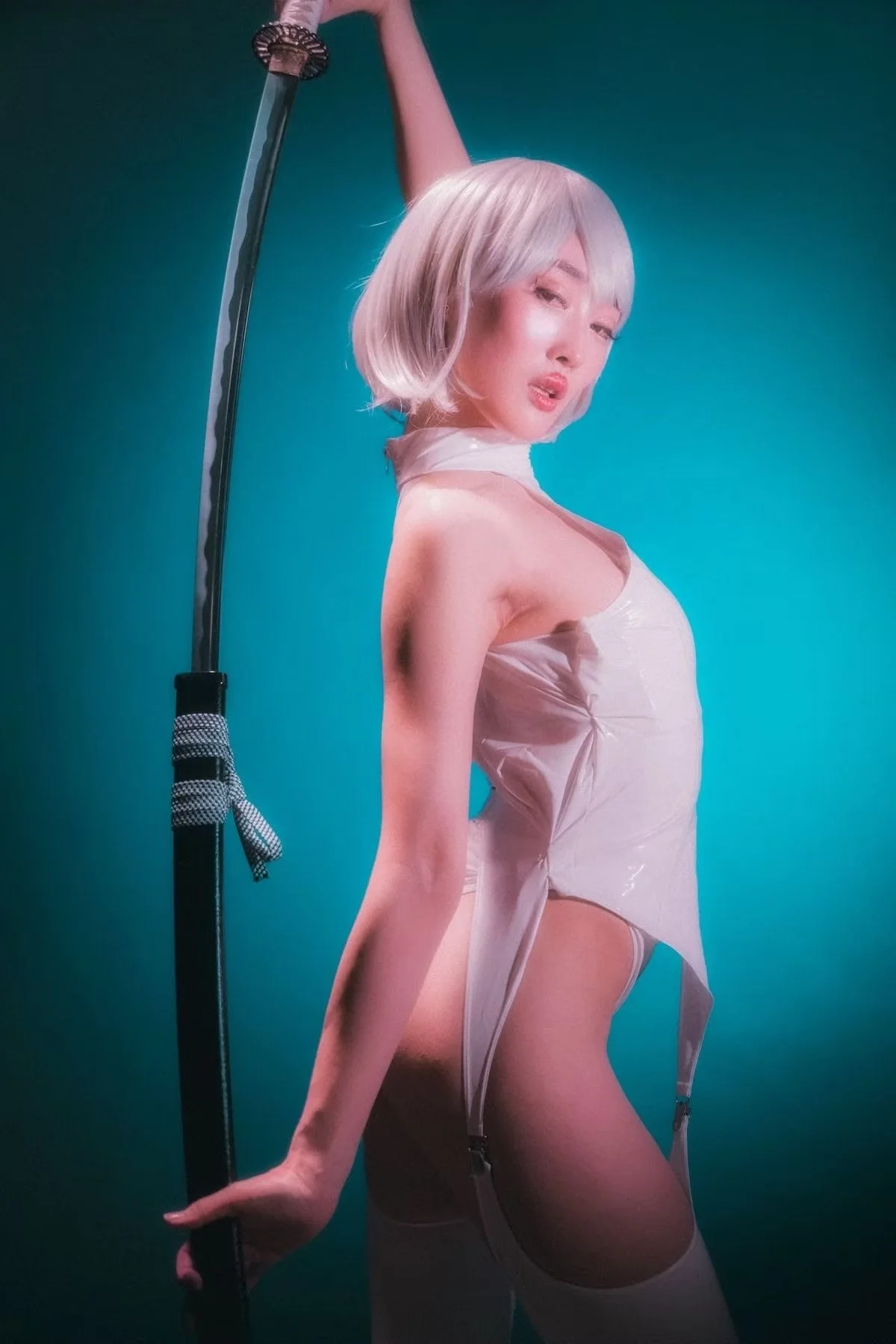 A woman with platinum blonde hair and light skin posing against a teal background. She wears a sheer, beige, high-slit dress with garter straps, holding a katana sword with a patterned grip.